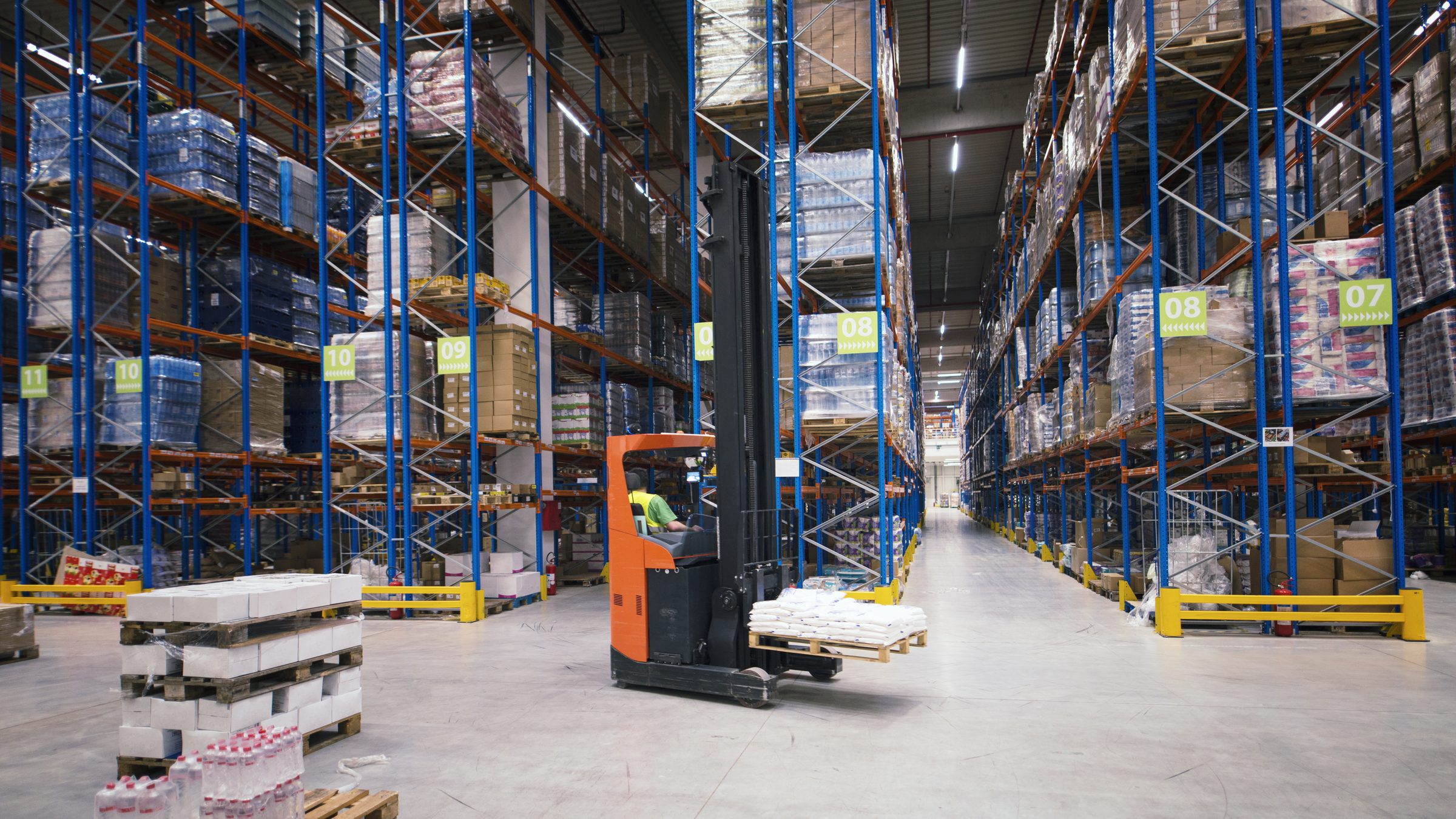 A distribution warehouse aisle, racking stacked with palletised stock, a reach truck lifting a pallet.