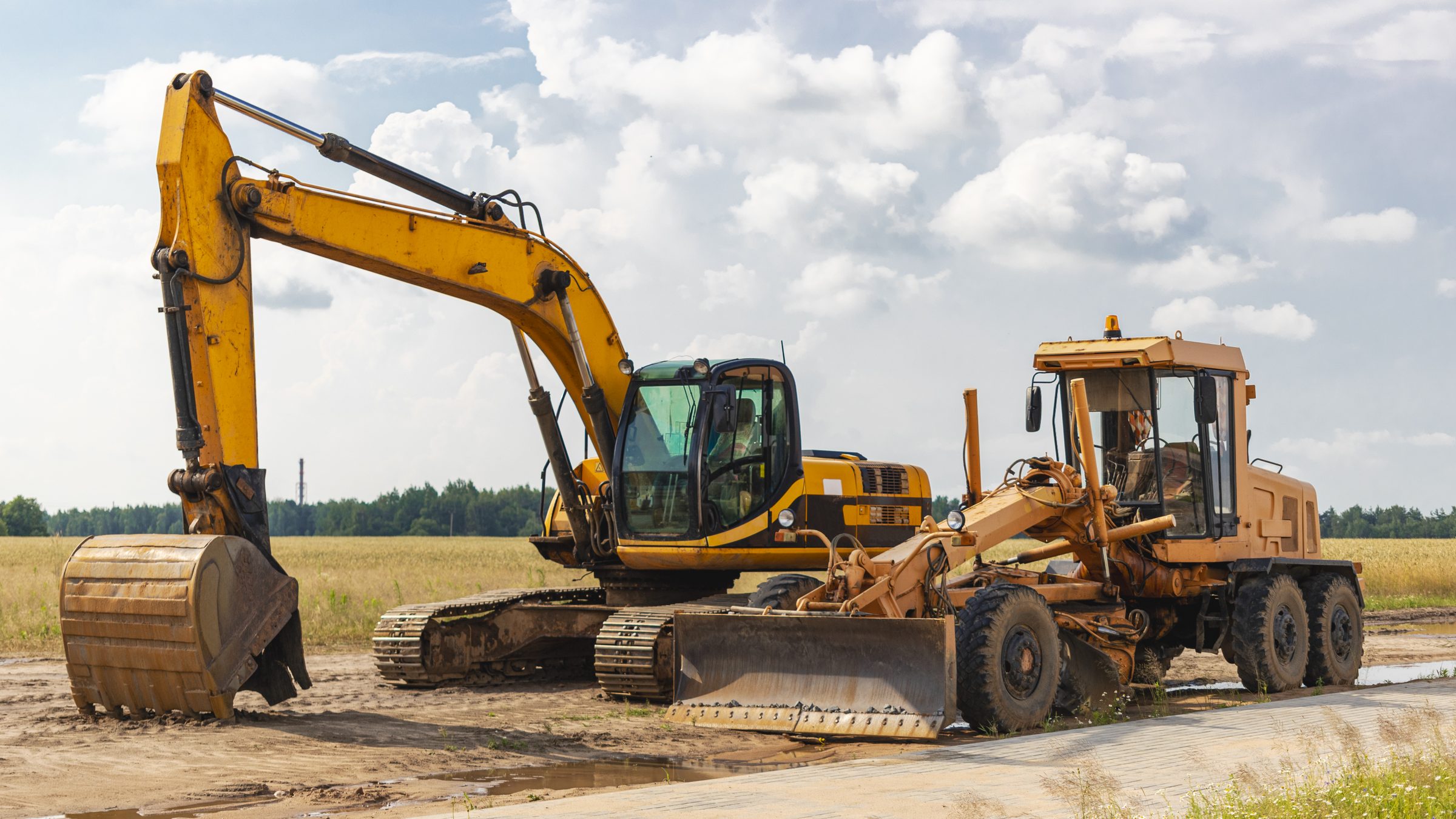 A tracked excavator and a motor grader parked side by side at a roadworks site, a field and treeline behind them.